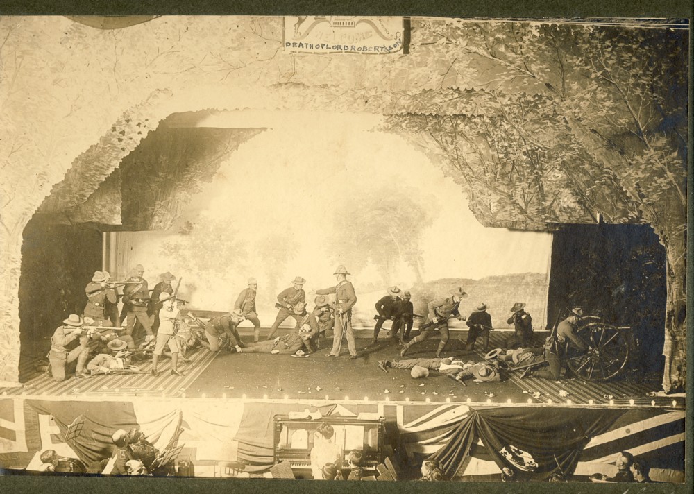Image for 'The Death of Lord Robert's Son'.  A photograph of an reenactment of Frederick Hugh Sherston Roberts death done on stage with military band in an orchestra pit in the foreground