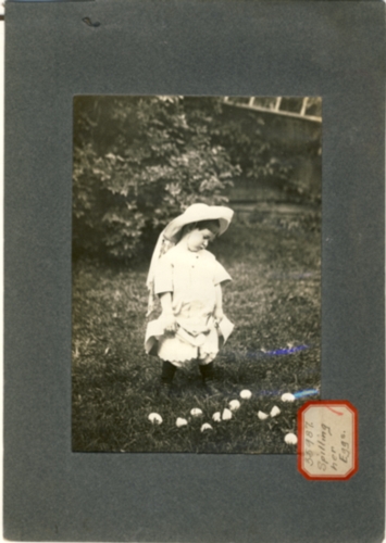 Image for Photograph of child with petticoats & hat looking down at a pile of eggs in the grass (Sticker reads Spilling Her Eggs)
